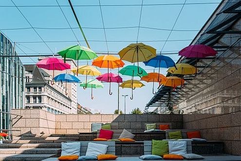 Bunte Regenschirme im Zeichen der Demokratie als Dekoration vor blauem Himmel über bunten Sitzkissen einer Gebäudetreppe..