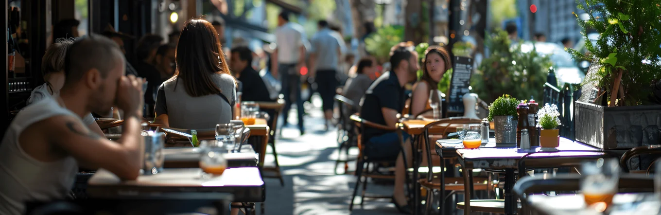 Straßencafé mit besetzten Tischen, Passanten und Baumreihe auf belebter innerstädtischer Straße