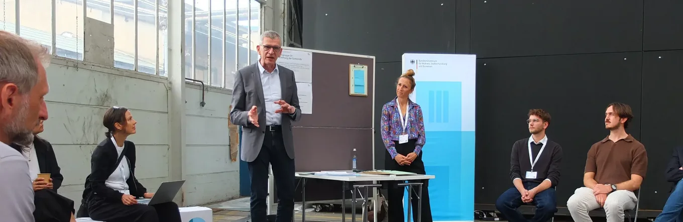 Menschen sitzen in einem Stuhlkreis und hören Bernhard Faller vom vhw zu, der an einer Tafel steht.