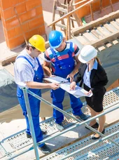 Weiterbildung Projektmanagerin Bauherren Diese Baustellenszene zeigt ein interdisziplinäres Team, bestehend aus Bauarbeitern und einer Architektin, die gemeinsam Baupläne auf einem Gerüst prüfen.