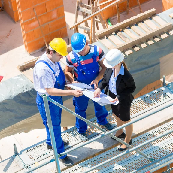 Weiterbildung Projektmanagerin Bauherren Diese Baustellenszene zeigt ein interdisziplinäres Team, bestehend aus Bauarbeitern und einer Architektin, die gemeinsam Baupläne auf einem Gerüst prüfen.