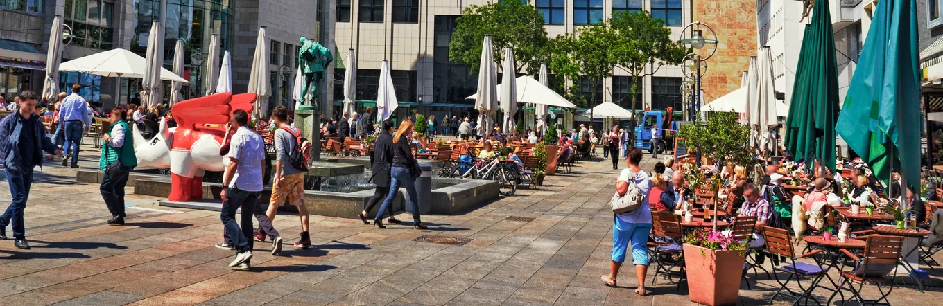Sonniger Platz in Dortmund mit Außengastronomie, Kunstwerk, Sonnenschirmen und vielen Besuchern zwischen modernen Gebäuden.