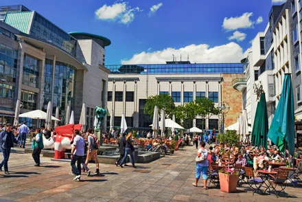 Sonniger Platz in Dortmund mit Außengastronomie, Kunstwerk, Sonnenschirmen und vielen Besuchern zwischen modernen Gebäuden.