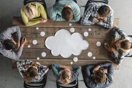 Acht Personen von oben um Holztisch mit großer weißer Cloud und kleinen Punkten als Symbol für Vernetzung.