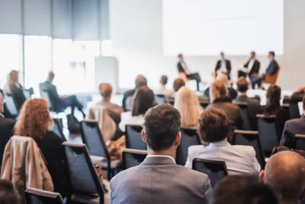 Publikum verfolgt Podiumsdiskussion mit vier Personen auf einer Bühne im Konferenzsaal
