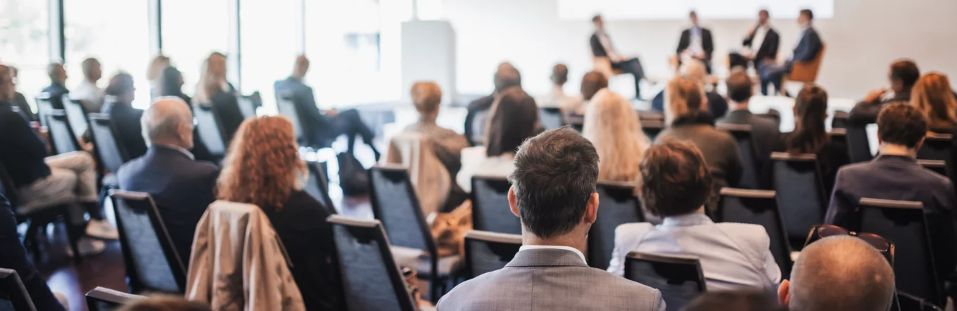 Publikum verfolgt Podiumsdiskussion mit vier Personen auf einer Bühne im Konferenzsaal