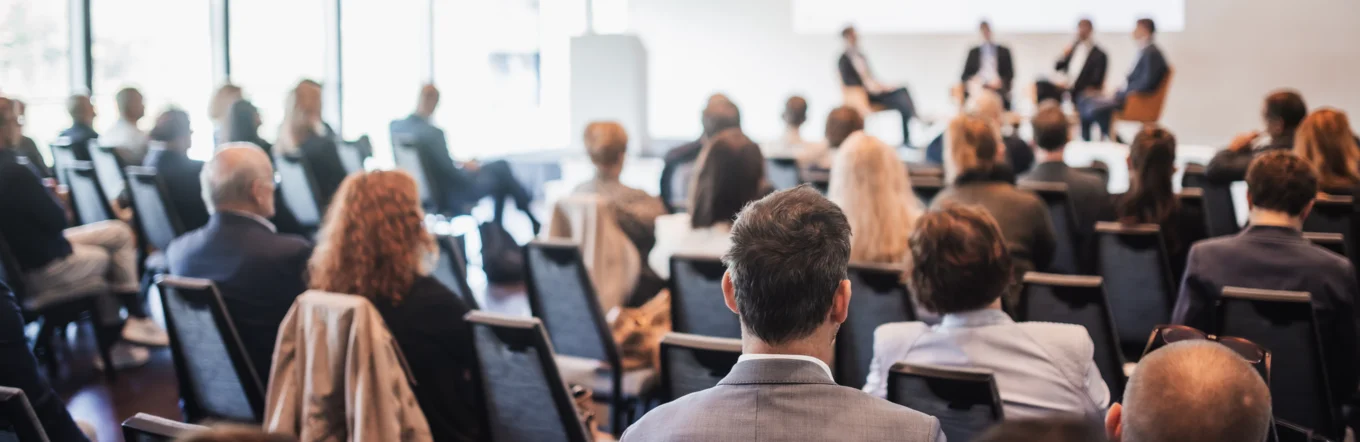 Publikum verfolgt Podiumsdiskussion mit vier Personen auf einer Bühne im Konferenzsaal