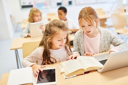 Foto: 2 Grundschülerinnen sitzenn einem Klassenraum sitzen an einem Laptop und schauen zusammen in ein Buch