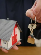 Basiswissen Grundbuchrecht Person hält einen Schlüssel in zur Kamera und hält in der anderen Hand ein Kleines Haus.