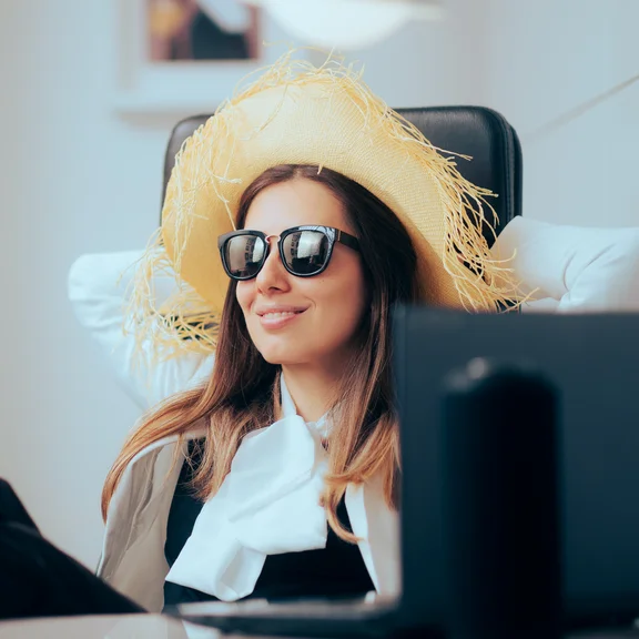 webinarsommer2026-c-nicoletaionescu-adobestock Frau mit Strohhut und Sonnenbrille sitzt zurückgelehnt vor Laptop im Büro, Füße auf dem Schreibtisch