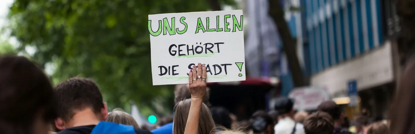 Demonstrantin mit Plakat „Uns allen gehört die Stadt" in Menschenmenge auf belebter innerstädtischer Straße.