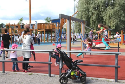 Öffentlicher Spielplatz mit Schaukelanlage, Klettergerüst, Familien und Kinderwagen an bewölktem Sommertag