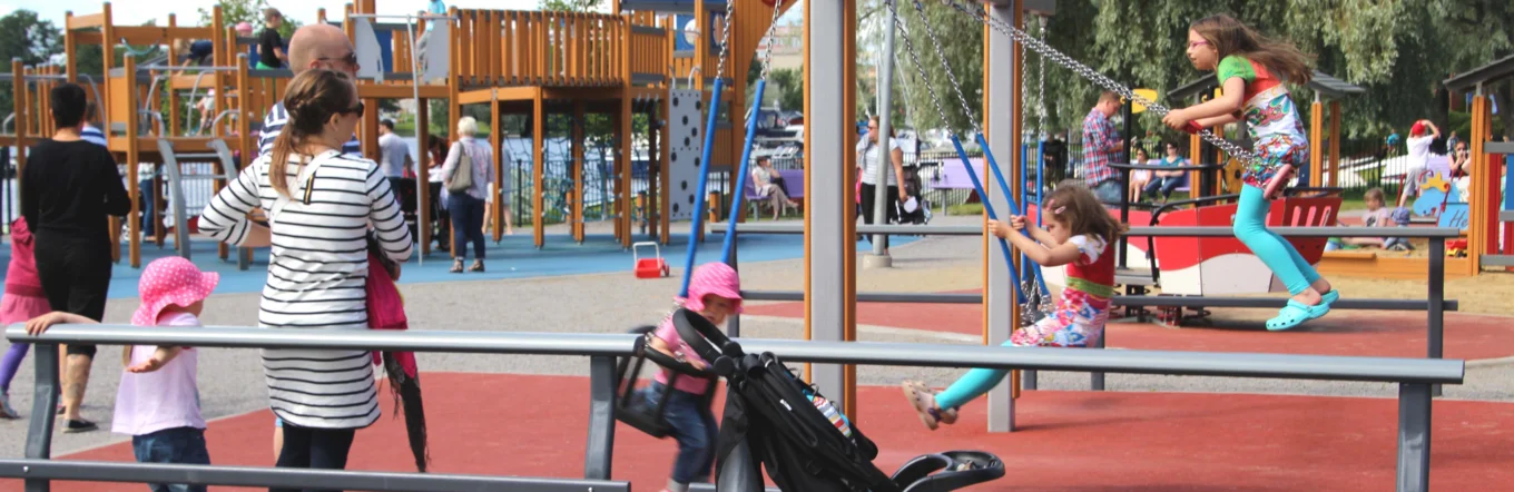Öffentlicher Spielplatz mit Schaukelanlage, Klettergerüst, Familien und Kinderwagen an bewölktem Sommertag