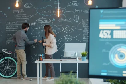 Zwei junge Menschen arbeiten gemeinsam kreativ an einer Tafelwand, im Vordergrund ist ein Computerbildschirm mit Zahlen und Grafiken zu sehen