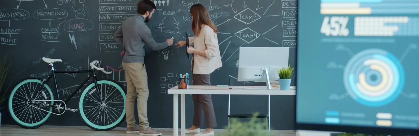 Zwei junge Menschen arbeiten gemeinsam kreativ an einer Tafelwand, im Vordergrund ist ein Computerbildschirm mit Zahlen und Grafiken zu sehen