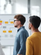 Zertifikatslehrgang Prozessmanager Das Bild zeigt eine Gruppe von Menschen, die vor einem Whiteboard mit einem Flussdiagramm oder Organisationsplan stehen.
