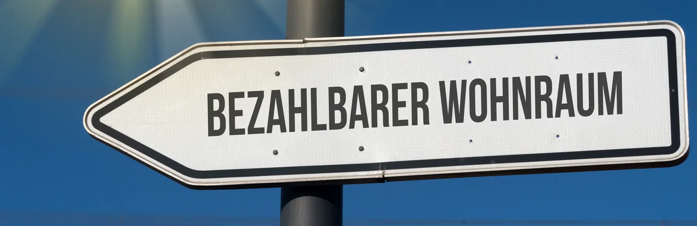 Wegweiserschild mit Aufschrift „Bezahlbarer Wohnraum" vor blauem Himmel mit Sonnenstrahlen.