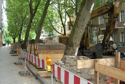 Straßenbaustelle mit Bagger und rot-weißen Absperrungen entlang einer Allee