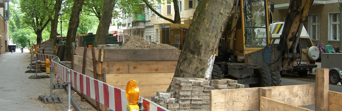 Straßenbaustelle mit Bagger und rot-weißen Absperrungen entlang einer Allee