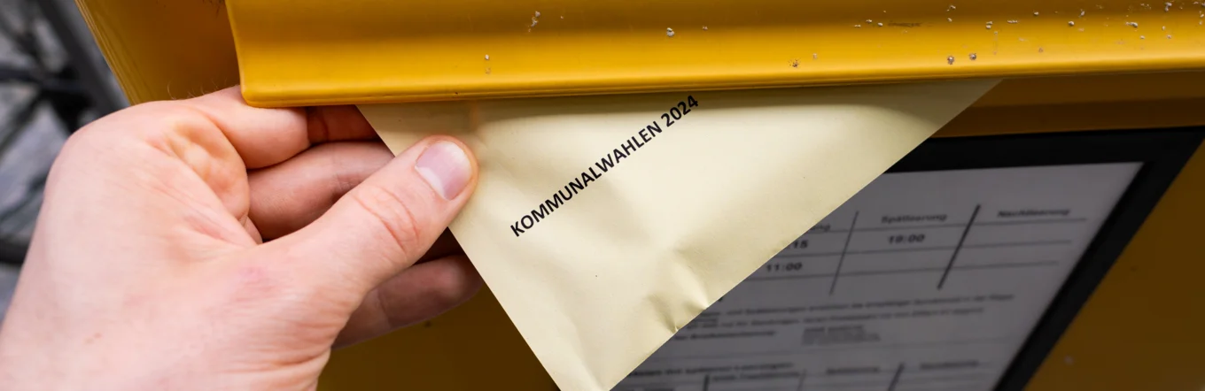Briefwahlumschlag „Kommunalwahlen 2024" wird in gelben Briefkasten eingeworfen
