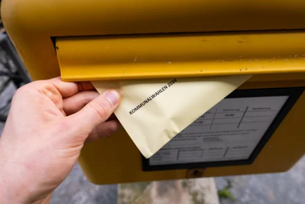 Briefwahlumschlag „Kommunalwahlen 2024" wird in gelben Briefkasten eingeworfen