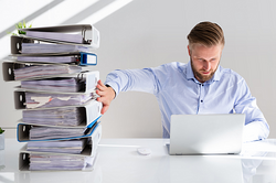 Foto symbolisiert die Digitalisierung der Verwaltung: Mitarbeiter schiebt Aktenberg zur Seite und arbeitet am Laptop