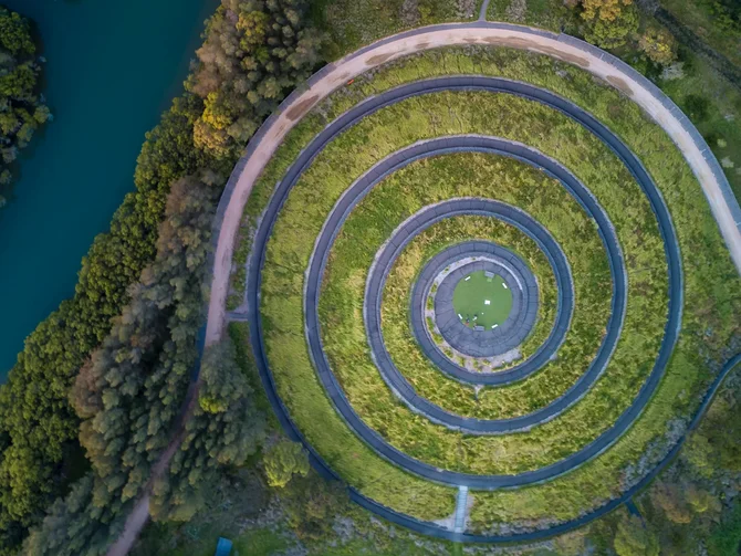 Umweltrecht und Klimaschutz Luftaufnahme einer spiralförmigen Grünanlage neben einem türkisfarbenen Fluss mit Brücke und Baumbestand
