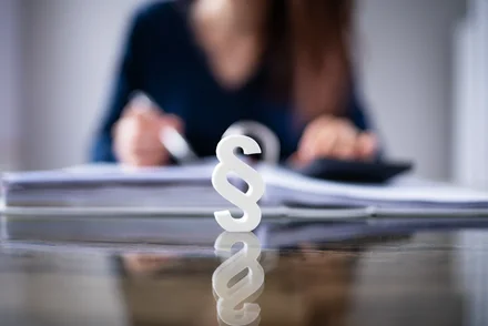 Weißes Paragraphensymbol im Fokus vor unscharfem Hintergrund mit Person an Schreibtisch und Dokumenten.