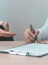 Grundlagenwissen Vergabewesen Zwei Personen führen ein Gespräch. Die rechts stehende Person ist kurz davor, ein Dokument zu unterschreiben. Im Vordergrund sind die Waage der Gerechtigkeit und ein Richterhammer zu sehen.