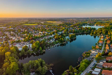  Panoramaluftbild einer norddeutschen Kleinstadt mit See, Wohnbebauung und Windrädern im Abendlicht
