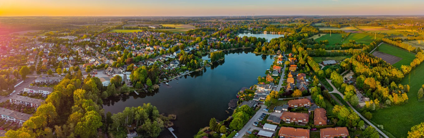 Panoramaluftbild einer norddeutschen Kleinstadt mit See, Wohnbebauung und Windrädern im Abendlicht