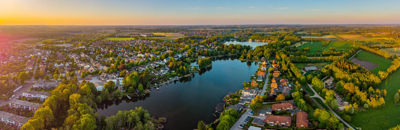 Panoramaluftbild einer norddeutschen Kleinstadt mit See, Wohnbebauung und Windrädern im Abendlicht
