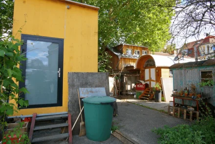Urbanes Wohnprojekt mit gelbem Kleingebäude, Bauwagen und gemeinschaftlich genutztem Außenbereich