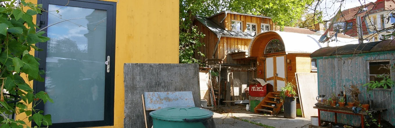 Urbanes Wohnprojekt mit gelbem Kleingebäude, Bauwagen und gemeinschaftlich genutztem Außenbereich