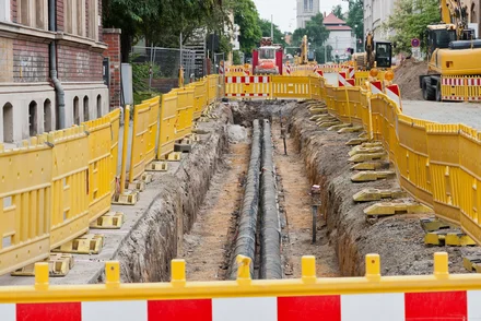 Offene Baugrube mit Fernwärmerohren zwischen historischen Häusern, gelben Bauzäunen und Baumaschinen.
