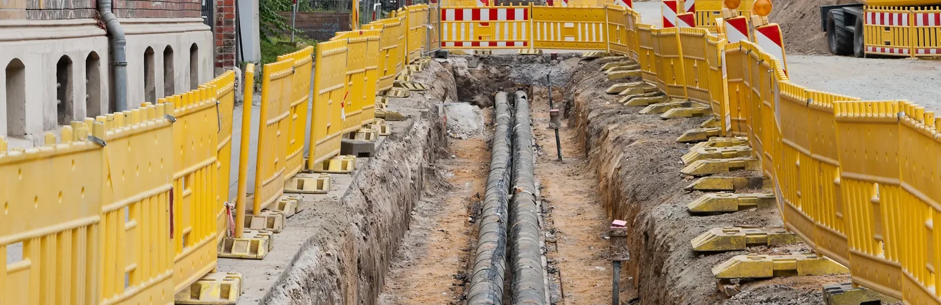 Offene Baugrube mit Fernwärmerohren zwischen historischen Häusern, gelben Bauzäunen und Baumaschinen.