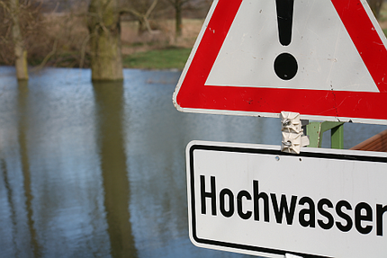 Hochwasser-Warnschild vor einem über die Ufer getretenen Fluss