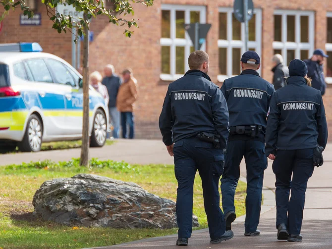polizei-ordnungsrecht-c-ronald-rampsch-adobestock Drei Ordnungsamtsbedienstete von hinten auf einem Gehweg, Polizeifahrzeug und Passanten im Hintergrund
