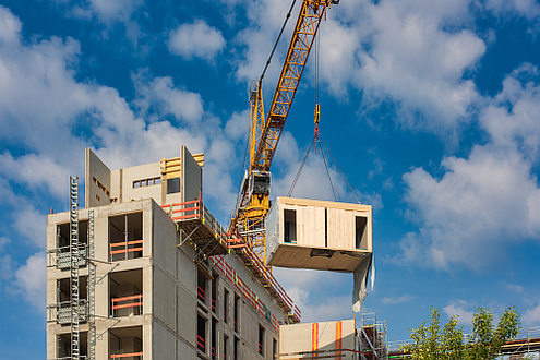 Foto eine Rohbaus in Modulbauweise. Ein Kran hebt ein Baumodul auf das Gebäude.