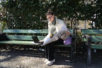 Eine Frau auf einer Parkbank liest Zeitung und arbeitet am Computer 