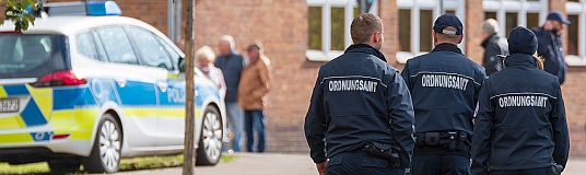 Foto: Polizei- und Ordnungsrecht-Mitarbeiter stehen auf der Straße vor einem Polizeiauto. 