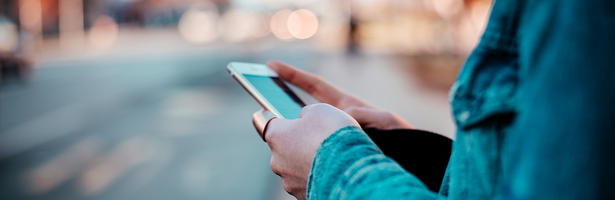 Person hat ein Smartphone in der Hand, im Hintergrund ist verschwommen eine urbane Umgebung zu erkennen